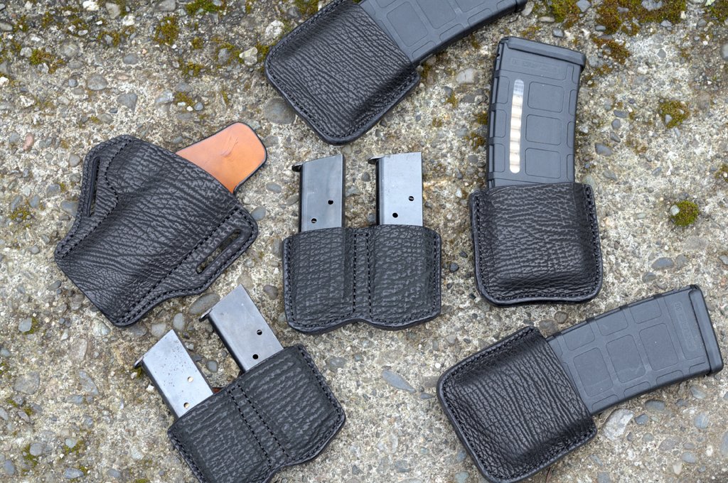 A photo of a set of holsters made from black sharkskin with cognac cowhide lining. The set includes a strong side holster for a 1911 pistol, two double magazine pouches for the pistol and three single magazine pouches for an AR-15 rifle. All are sitting on a mossy cement surface