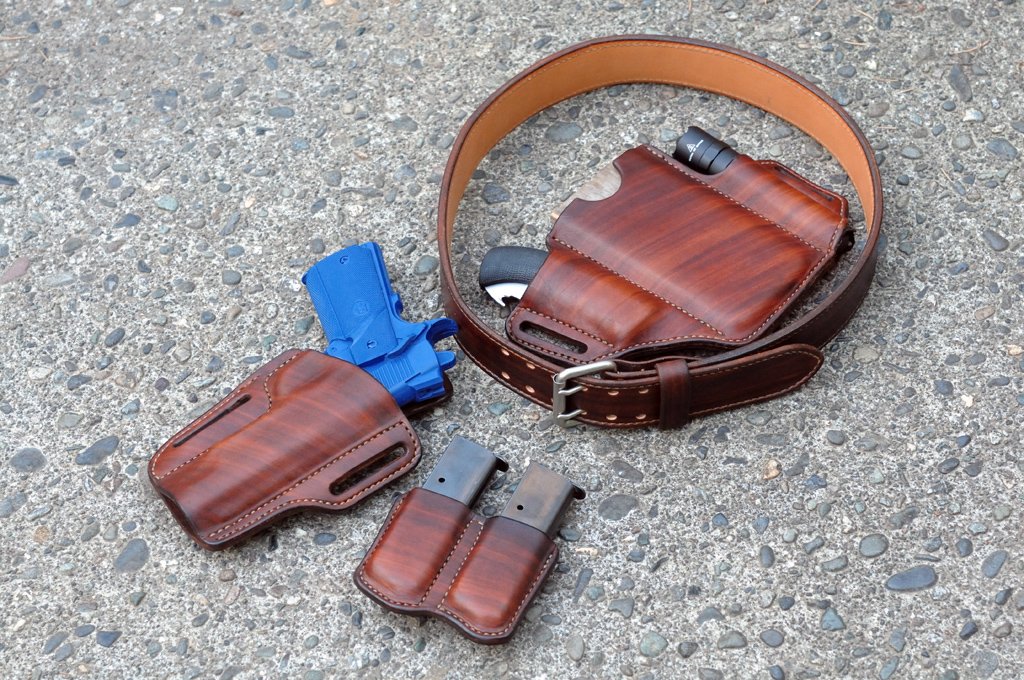 Artistic view of a matching set of leather holsters for a 1911 pistol, double magazine pouch, combo holster for a folding knife, phone and flashlight, and a belt.