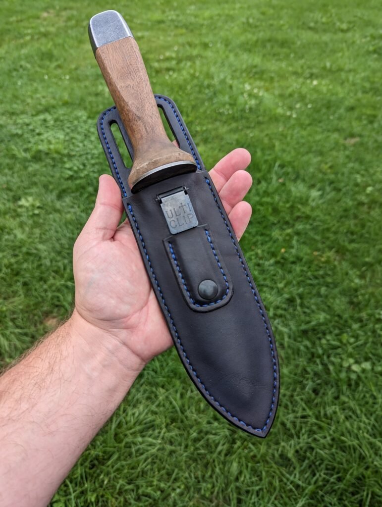Front view of black leather boot sheath for Hori Hori Japanese gardening knife held in front of green grass background.