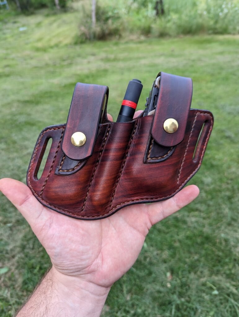 Front view of a sheath for a Leatherman OHT multitool, Coast flashlight and folding Buck knife. The sheath is made from hand dyed brown leather and is shown held in front of a grassy backdrop.