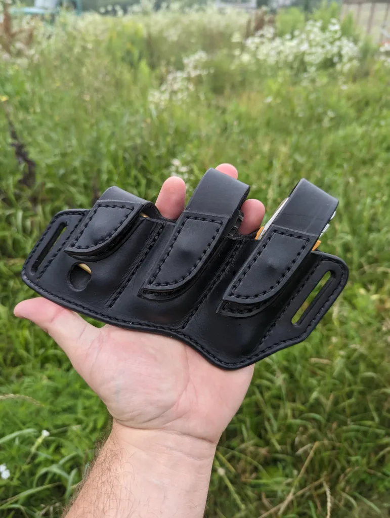 Front view of a sheath for a Leatherman Signal with driver bit set and a Zippo lighter. The sheath is made from black leather and is held in front of green grass.