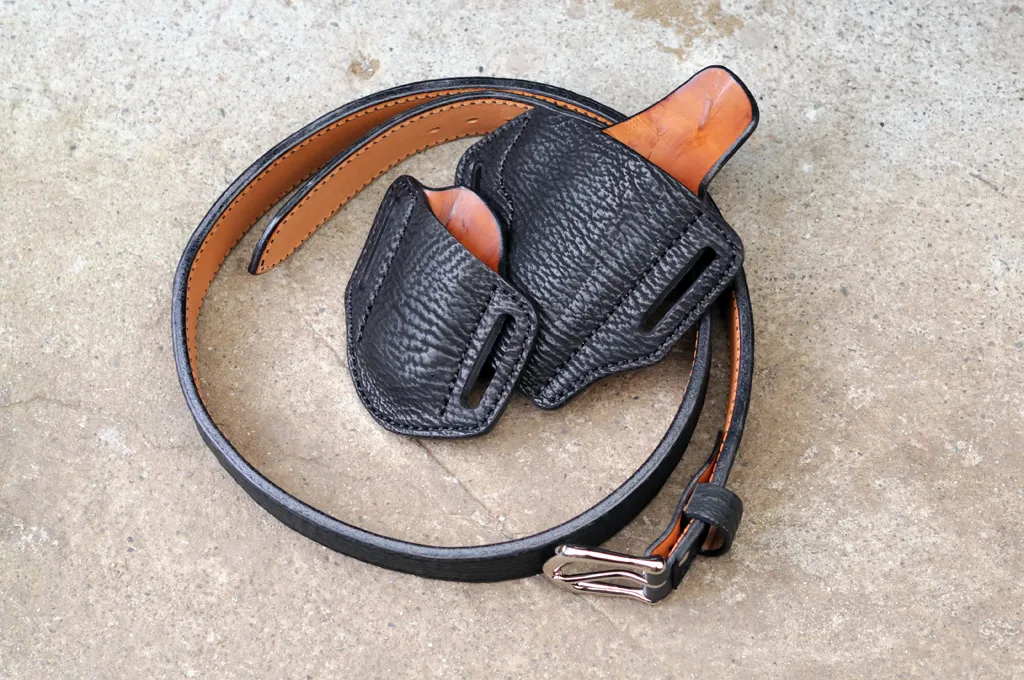 Artistic photo of a holster, magazine pouch and belt made of black sharkskin for a 1911 pistol. The set is sitting on a cement surface