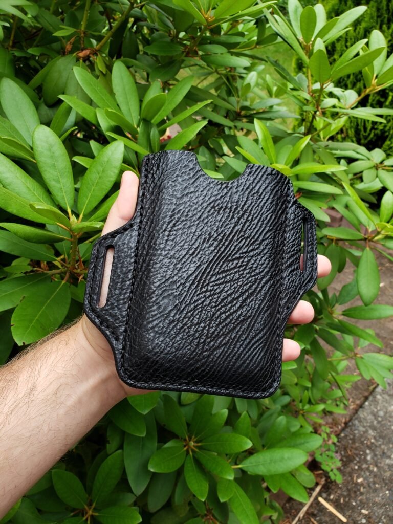 Front view of a phone holster made from black sharkskin and cowhide leather held in front of a rhododendron plant.