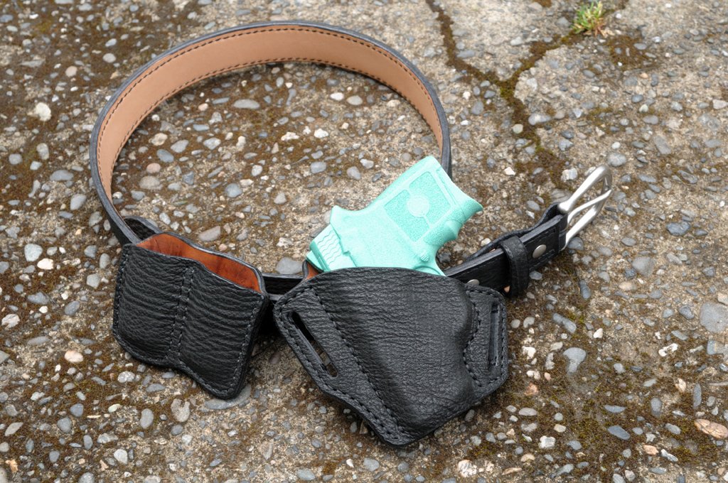 Artistic photo of a holster, magazine and belt set for a Smith and Wesson Bodyguard pistol sitting on a mossy cement surface. The set is made from black sharkskin and light brown cowhide leather.