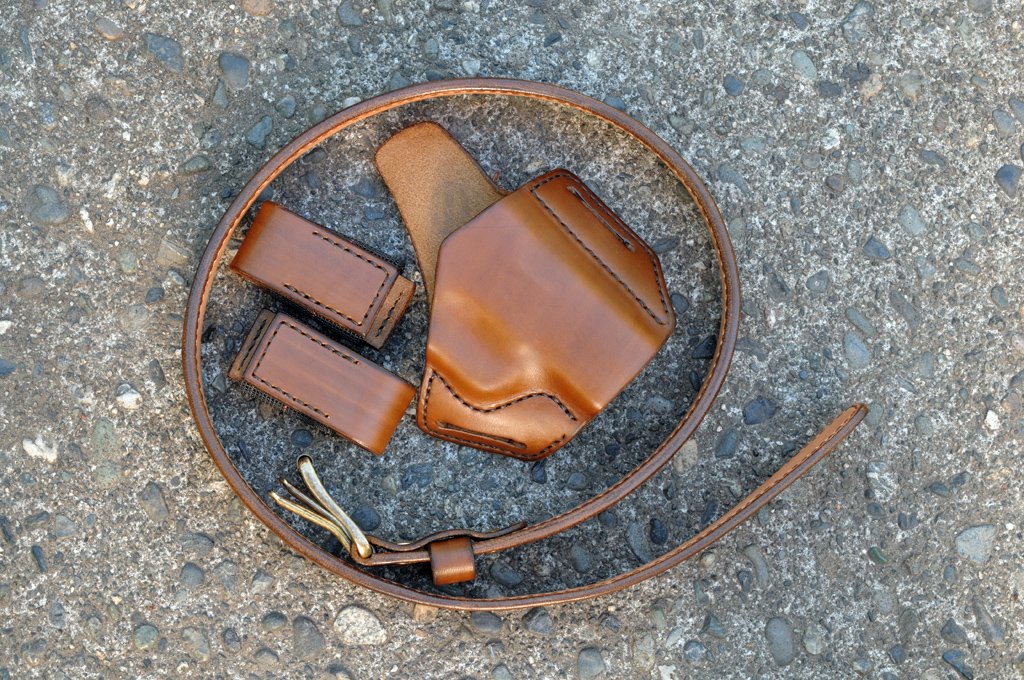 A photo of a holster, magazine pouches and belt set for a Walther PPS pistol. The set is made from matching medium brown leather and sitting on a cement surface.