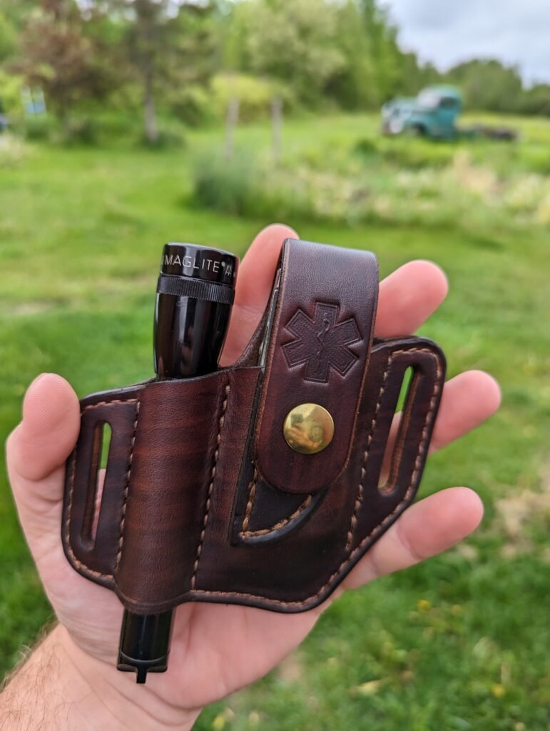 Front view of a sheath for a Leatherman multitool and Maglite flashlight. It has the Emergency Medical Services emblem stamped on the front and is made from chocolate brown dyed leather