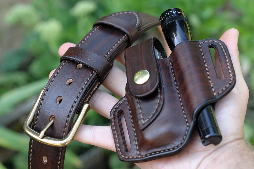Artistic photo of a leather belt and matching sheath for a Leatherman multitool and Maglite flashlight held in a hand with green vegetation in the background