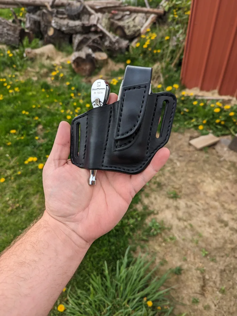 Front view of a black leather sheath for a Leatherman multitool and small ratchet. It's held in a hand with green grass and dandelions in the background.
