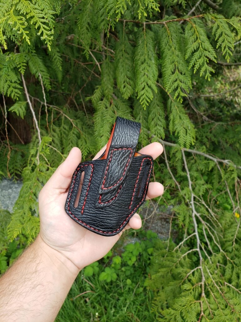 Front view of a sheath for a pair of Knipex pliers made from black sharkskin with red stitching held in a hand with green vegetation in the background