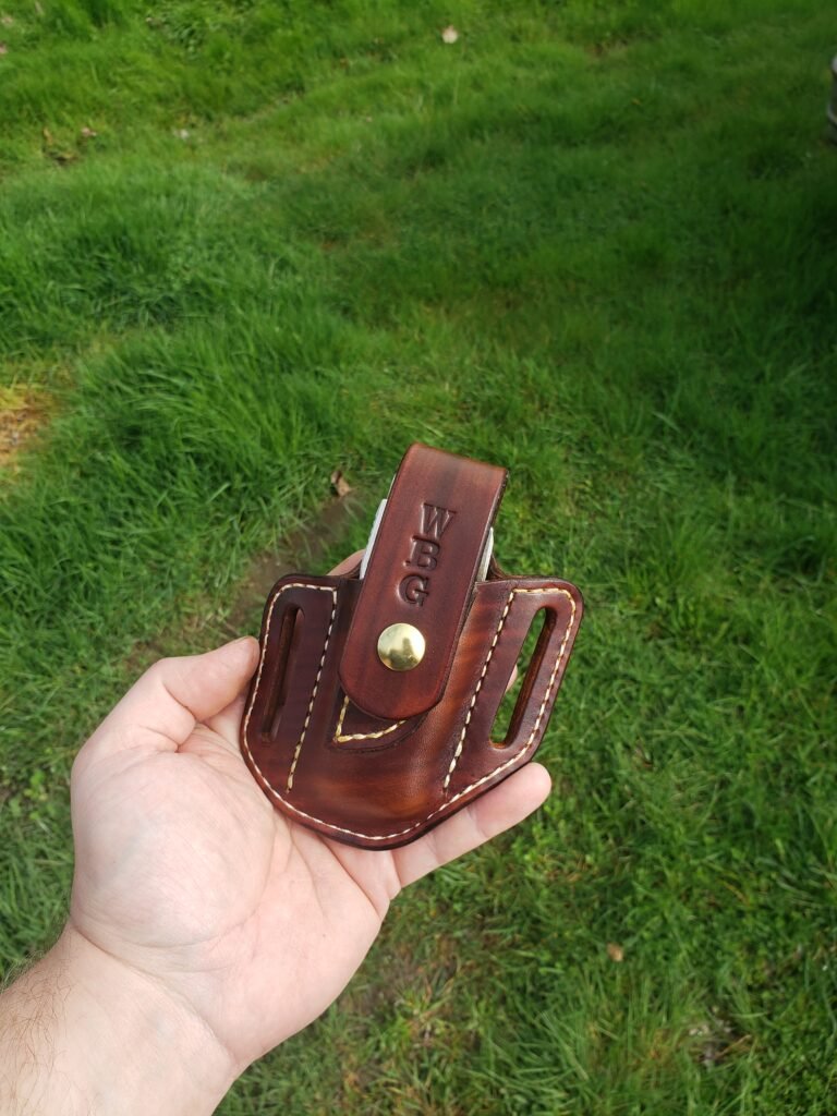 Front view of a sheath for a Leatherman Surge made from brown leather with white thread with the letters WBG stamped on the front. The sheath is held in a hand with green grass in the background